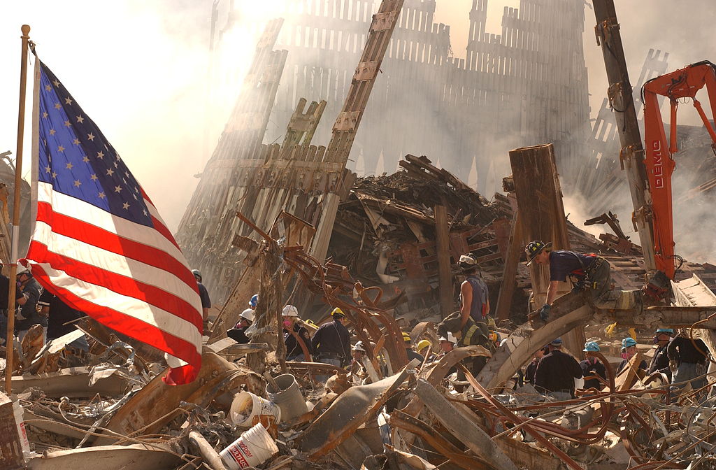 Rescue workers climb over and dig through piles of rubble from the destroyed World Trade Center as the American flag flies over the debris. (Andrea Booher/Wikimedia Commons)